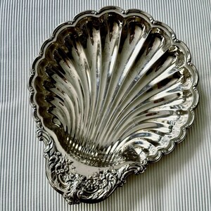 K&ouml;nnte beinhalten: Eine polierte silberfarbene Metallschale in Form einer gro&szlig;en Jakobsmuschel. Das Innere weist tiefe, strahlenf&ouml;rmige Rillen auf, w&auml;hrend die Basis mit detaillierten, erhabenen Blumen- und Schn&ouml;rkelverzierungen entlang des Randes versehen ist.