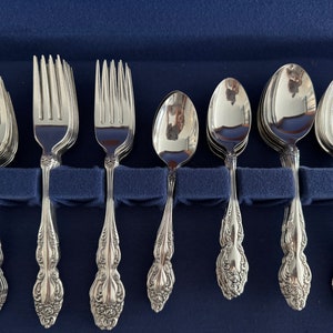 May include: A set of ornate silverware, including forks and spoons, arranged in a blue case. The silverware has a detailed floral pattern on the handles and a shiny, reflective surface. The set appears to be for formal dining.