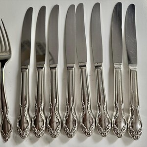 May include: A collection of stainless steel silverware, including forks and knives. The knives have a decorative handle design. The silverware is arranged on a white surface, suggesting a formal dining setting.