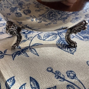 May include: A polished silver bowl with a reflective surface and ornate, curved feet. The bowl features a smooth, rounded body that mirrors the blue and white floral pattern of the fabric underneath it, creating a clear visual reflection.