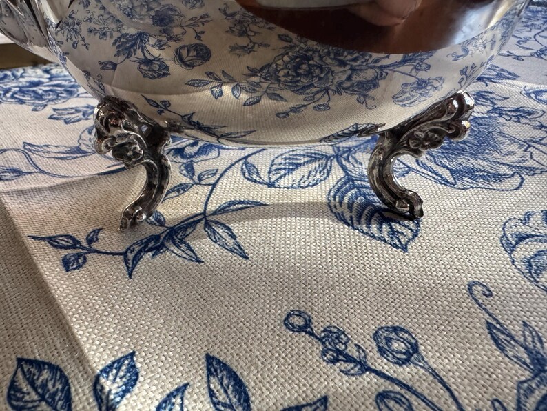 May include: A polished silver bowl with a reflective surface and ornate, curved feet. The bowl features a smooth, rounded body that mirrors the blue and white floral pattern of the fabric underneath it, creating a clear visual reflection.