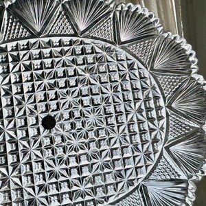 May include: Close-up of a clear, cut-glass serving plate. The plate features a detailed geometric pattern with starburst and fan-shaped designs. The rim has a scalloped edge, adding to its decorative appeal. A small hole is in the center.