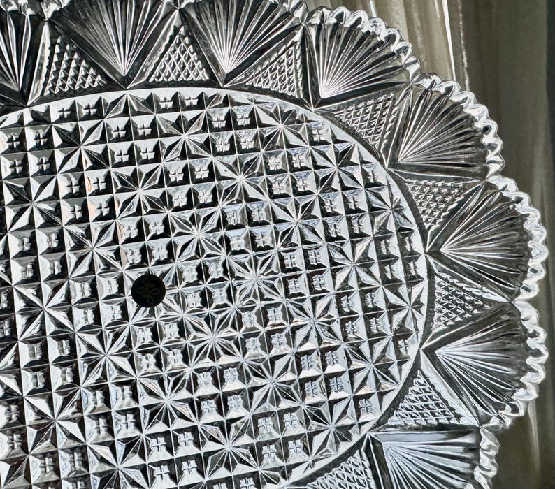 May include: Close-up of a clear, cut-glass serving plate. The plate features a detailed geometric pattern with starburst and fan-shaped designs. The rim has a scalloped edge, adding to its decorative appeal. A small hole is in the center.