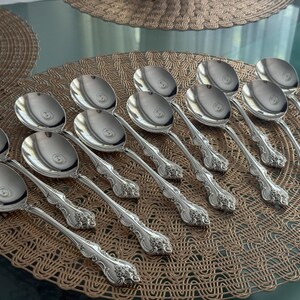May include: A collection of silver soup spoons arranged on a woven placemat. The spoons have ornate, detailed handles and large, rounded bowls. The spoons are reflective, and the arrangement suggests a formal dining setting.