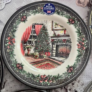 May include: A Royal Stafford plate with a Christmas scene. The plate features a black rim with a dotted pattern and floral accents. The central image depicts a decorated Christmas tree, fireplace, and red curtains.