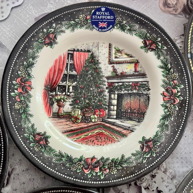 May include: A Royal Stafford plate with a Christmas scene. The plate features a black rim with a dotted pattern and floral accents. The central image depicts a decorated Christmas tree, fireplace, and red curtains.