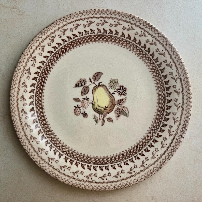 May include: A round ceramic plate with a cream-colored base featuring a central illustration of a yellow pear surrounded by brown leaves and small flowers. The rim is decorated with a repeating brown geometric and floral pattern.