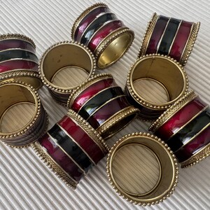 May include: A set of napkin rings. The rings have a gold-coloured metal base, with bands of red and black enamel. The edges are decorated with small, gold-coloured beads. These rings would be a stylish addition to any dining table.