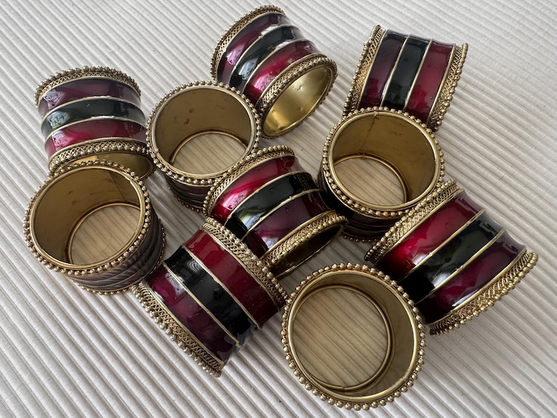 May include: A set of napkin rings. The rings have a gold-coloured metal base, with bands of red and black enamel. The edges are decorated with small, gold-coloured beads. These rings would be a stylish addition to any dining table.