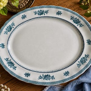 May include: An oval ceramic serving platter with a white base and a scalloped edge. The rim features a dark teal floral pattern interspersed with small bouquets and a delicate geometric border line that encircles the central serving area.