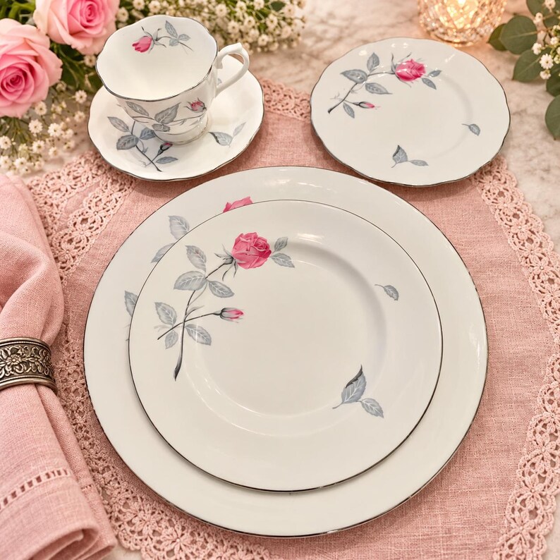 May include: A white porcelain dinnerware set featuring a delicate pink rose and grey leaf pattern with silver-toned rims. The set includes a teacup on a saucer, a small plate, a salad plate, and a large dinner plate, all with scalloped edges.