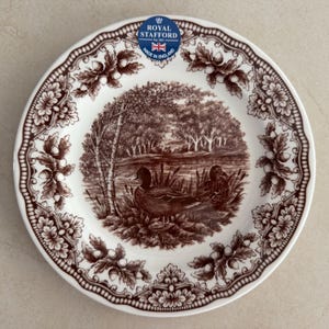 Op de afbeelding: Een wit keramieken bord met een bruine transferprint van twee eenden bij een vijver. De rand is versierd met een herhalend patroon van eikenbladeren en eikels. Een blauwe sticker vermeldt Royal Stafford Est 1845 Made in England.