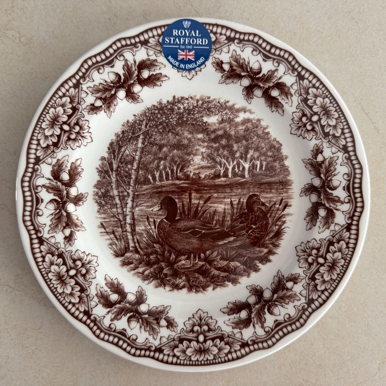 Op de afbeelding: Een wit keramieken bord met een bruine transferprint van twee eenden bij een vijver. De rand is versierd met een herhalend patroon van eikenbladeren en eikels. Een blauwe sticker vermeldt Royal Stafford Est 1845 Made in England.