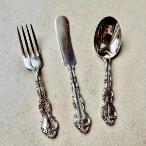 May include: A set of silver cutlery comprising a fork, butter knife, and spoon. Each piece has an ornate handle design. The fork has four prongs, the knife has a rounded blade, and the spoon has a deep bowl. The set is arranged on a neutral-toned surface.