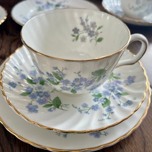 May include: A white teacup, saucer, and dessert plate set with a delicate blue floral pattern and gold trim. The teacup is centered on the saucer, with a matching dessert plate below. The set is on a wooden surface.