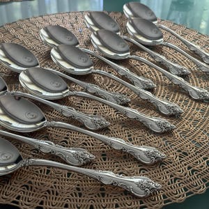 May include: A set of silver spoons with decorative handles, displayed on a woven placemat. The spoons have a polished surface, and the handles feature a detailed floral pattern. The spoons are designed for serving.