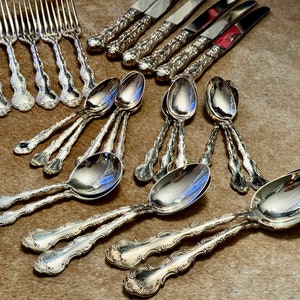 May include: A set of silverware, including forks, knives, and spoons, with ornate, silver-plated handles. The silverware is arranged on a brown, textured surface.