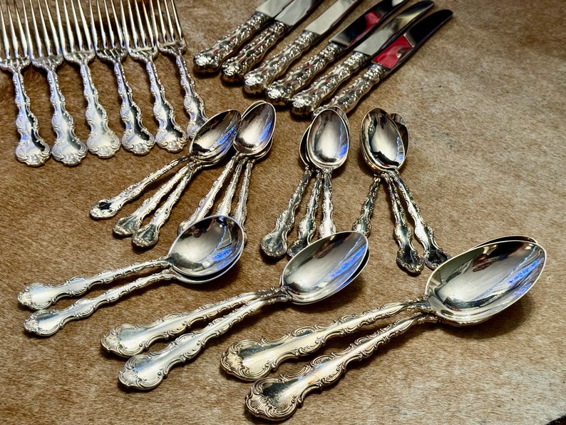 May include: A set of silverware, including forks, knives, and spoons, with ornate, silver-plated handles. The silverware is arranged on a brown, textured surface.