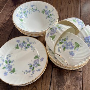 May include: A set of white porcelain dishes with a floral pattern of blue flowers and green leaves. The set includes bowls, plates, teacups, and saucers with gold trim. The dishes are arranged on a wooden surface.