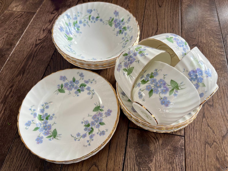 May include: A set of white porcelain dishes with a floral pattern of blue flowers and green leaves. The set includes bowls, plates, teacups, and saucers with gold trim. The dishes are arranged on a wooden surface.