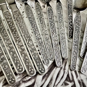 Puede incluir: Una colecci&oacute;n de cubiertos de metal plateado con un patr&oacute;n floral en relieve en los mangos. El dise&ntilde;o incluye representaciones detalladas de peque&ntilde;as flores, hojas y detalles en forma de estrella a lo largo de cada mango, con cuencos y hojas lisos.