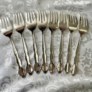 May include: A collection of eight silver forks arranged on a white, patterned tablecloth. The forks have ornate handles and tines, reflecting light. The silverware is likely for formal dining.