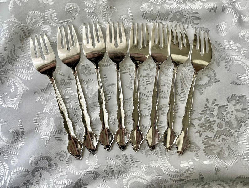 May include: A collection of eight silver forks arranged on a white, patterned tablecloth. The forks have ornate handles and tines, reflecting light. The silverware is likely for formal dining.