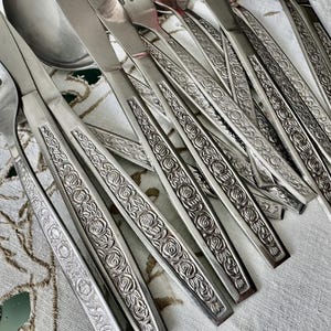 May include: A selection of stainless steel cutlery, including forks, knives, and spoons. The cutlery has a floral rose design on the handles. The utensils are arranged on a patterned tablecloth.