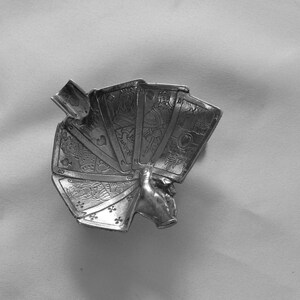 K&ouml;nnte beinhalten: Ein silberfarbener Aschenbecher in Form einer Hand, die ein F&auml;cher von Spielkarten h&auml;lt. Die Karten zeigen detaillierte Symbole und Zahlen. Der Aschenbecher hat eine kleine, rechteckige Ablage f&uuml;r Zigaretten.