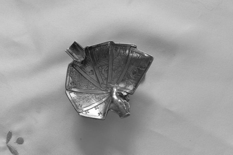 K&ouml;nnte beinhalten: Ein silberfarbener Aschenbecher in Form einer Hand, die ein F&auml;cher von Spielkarten h&auml;lt. Die Karten zeigen detaillierte Symbole und Zahlen. Der Aschenbecher hat eine kleine, rechteckige Ablage f&uuml;r Zigaretten.