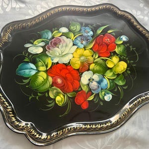 May include: Black decorative tray with a floral design. The tray features a scalloped edge with gold trim. The floral arrangement includes red, white, yellow, and blue flowers with green leaves.