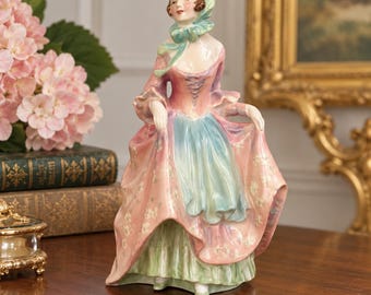 May include: A porcelain figurine of a person wearing a pink and blue gown with a matching headscarf. The figure features a fitted bodice with lace-up detailing, a full skirt with floral accents, and a ruffled green underskirt, standing in a pose.