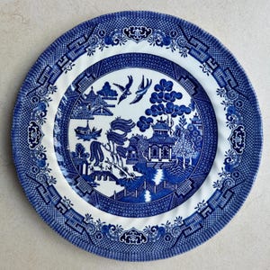 May include: A round ceramic dinner plate featuring a blue and white willow pattern. The central design depicts a landscape with a pagoda, a bridge, a boat, and two birds in flight. The wide rim is decorated with a geometric lattice border.