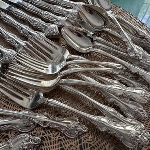 May include: An assortment of ornate silver cutlery, including forks, spoons, and knives, arranged on a decorative surface. The silverware features detailed floral patterns on the handles, reflecting light and creating a luxurious aesthetic. The collection suggests a formal dining setting.