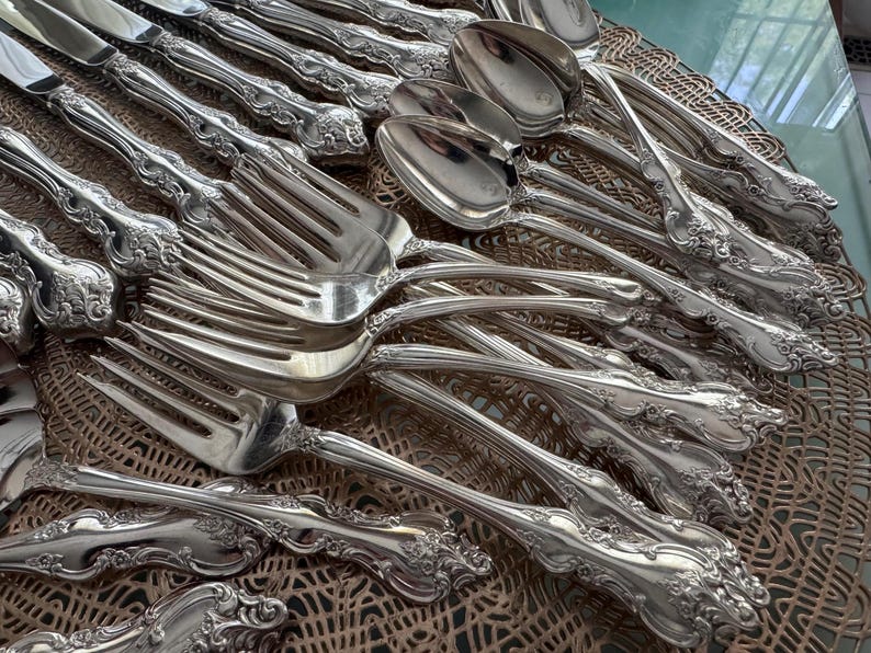 May include: An assortment of ornate silver cutlery, including forks, spoons, and knives, arranged on a decorative surface. The silverware features detailed floral patterns on the handles, reflecting light and creating a luxurious aesthetic. The collection suggests a formal dining setting.