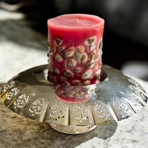 May include: A red pillar candle with embedded stones sits in a silver, scalloped-edge candle holder. The candle has a smooth top and a textured, stone-like exterior. The candle holder has an ornate design.