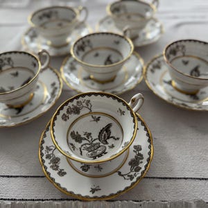 May include: A set of six white teacups and saucers with black and gold floral designs. The cups and saucers have a delicate, elegant look.