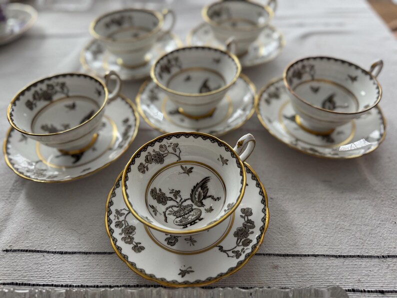 May include: A set of six white teacups and saucers with black and gold floral designs. The cups and saucers have a delicate, elegant look.
