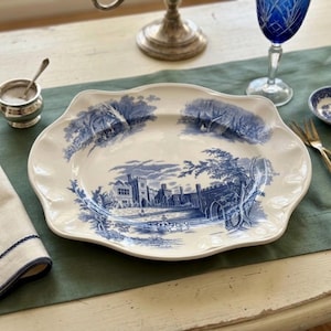 Op de afbeelding: Een grote, cr&egrave;mekleurige keramische serveerschaal met een geschulpte rand, voorzien van een gedetailleerde blauwe transferprint van een kasteel, bomen en een landschap. De schaal heeft een glanzende afwerking en een brede, ondiepe, ovale vorm.
