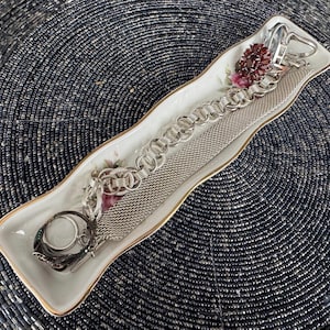 May include: A white porcelain dish with a floral design, holding two silver chains and a silver ring. The dish has a wavy edge and is on a black background.