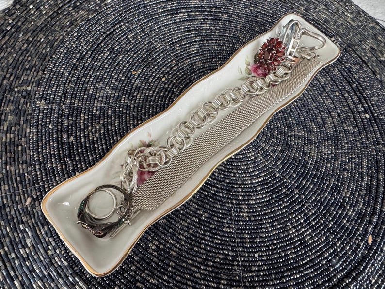 May include: A white porcelain dish with a floral design, holding two silver chains and a silver ring. The dish has a wavy edge and is on a black background.