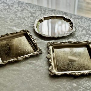 Puede incluir: Dos bandejas cuadradas de metal plateado con bordes florales en relieve y detalles grabados en el centro, acompa&ntilde;adas de una bandeja redonda de metal plateado con una superficie lisa y reflectante y un borde festoneado.