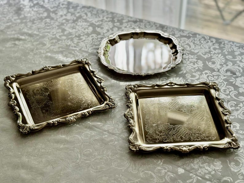 Puede incluir: Dos bandejas cuadradas de metal plateado con bordes florales en relieve y detalles grabados en el centro, acompa&ntilde;adas de una bandeja redonda de metal plateado con una superficie lisa y reflectante y un borde festoneado.