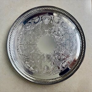 May include: A round silver-colored metal serving tray featuring an intricate embossed floral and scroll pattern across the center surface and a decorative pierced lattice border along the outer rim of the circular plate.
