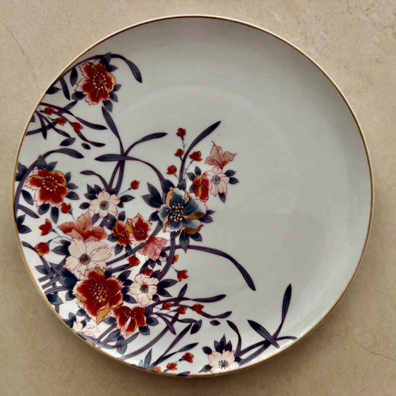Pu&ograve; includere: Un piatto rotondo in ceramica bianca con un delicato motivo floreale sul lato sinistro. Il design presenta fiori rosso scuro, rosa tenue e blu scuro con steli e foglie sottili. Il bordo del piatto &egrave; rifinito con un sottile bordo dorato.