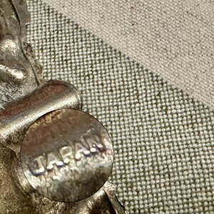 May include: A silver-toned metal earring featuring a textured, ornate design with a circular component clearly stamped with the word JAPAN. The metal surface shows signs of age and oxidation, with a metallic sheen and visible relief detailing.