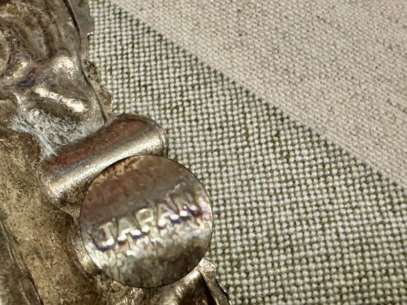 May include: A silver-toned metal earring featuring a textured, ornate design with a circular component clearly stamped with the word JAPAN. The metal surface shows signs of age and oxidation, with a metallic sheen and visible relief detailing.