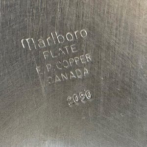 Puede incluir: Primer plano de una superficie met&aacute;lica plateada con el texto en relieve "Marlboro PLATE F.P. COPPER CANADA 2050". La superficie tiene una textura cepillada.