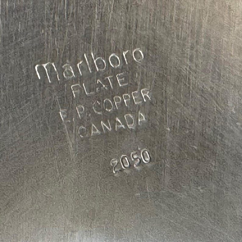 Puede incluir: Primer plano de una superficie met&aacute;lica plateada con el texto en relieve "Marlboro PLATE F.P. COPPER CANADA 2050". La superficie tiene una textura cepillada.