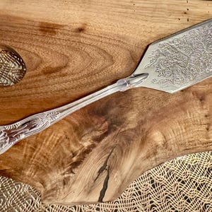 K&ouml;nnte beinhalten: Ein Tortenheber aus silberfarbenem Metall mit einem verzierten, gepr&auml;gten Blumen- und Rankenmuster auf der flachen Klinge. Der Griff ist schlank und weist an der Basis ein dekoratives Muschelmotiv sowie feine Details am Stiel auf.